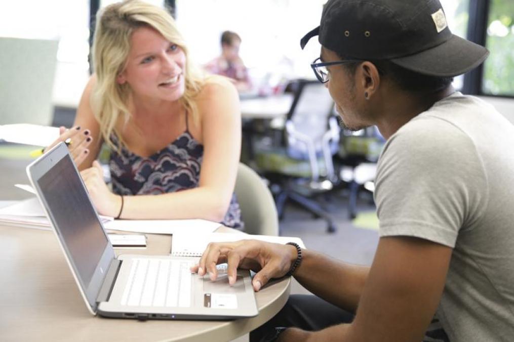 two students sitting next to each other talking around a lap top