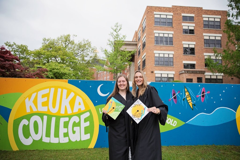 Sydnie Brown and Sarah Tower in graduation gowns in front of mural in their graduation robes 