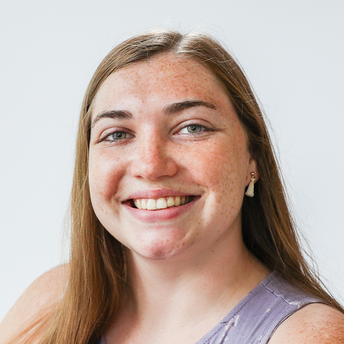 Smiling woman with long brown hair and freckles against a light background.
